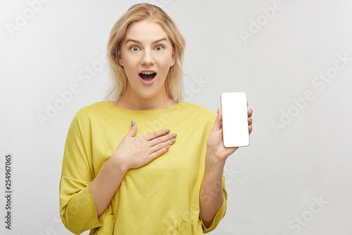 Shocked European blonde woman with mobile phone in hand, eyes bulging in surprise, emotions from what she sees on screen and keeps jaw dropped. Portrait of a beautiful girl isolated in white studio