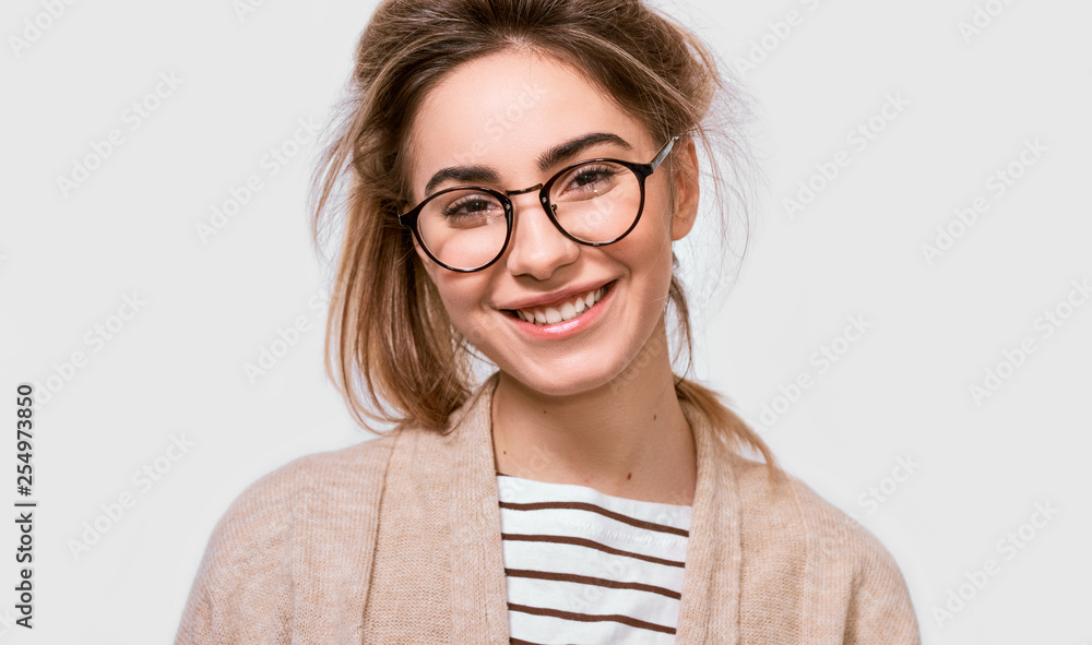 Close up portrait of beautiful positive female dressed in casual outfit, wearing round transparent eyeglasses with pleasant smile, looking to the camera and posing over white wall. People emotions