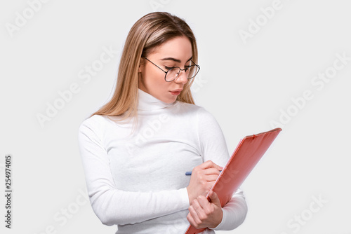 Serious young woman wearing white blouse and round transparent eyeglasses with red folder in hands, writing some notices. European female office worker posing on white studio background. Business