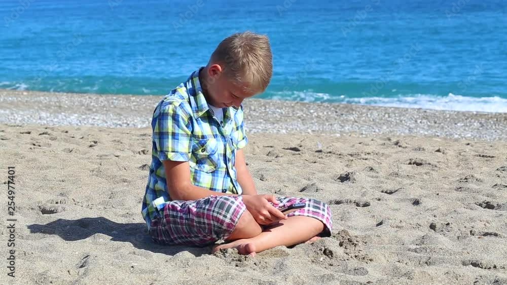 Cute white kid using smartphone while sitting on sand of summer sunny beach. Real time full hd video footage. Cute white kid using smartphone while sitting on sand of summer sunny beach. Real time full hd video footage.