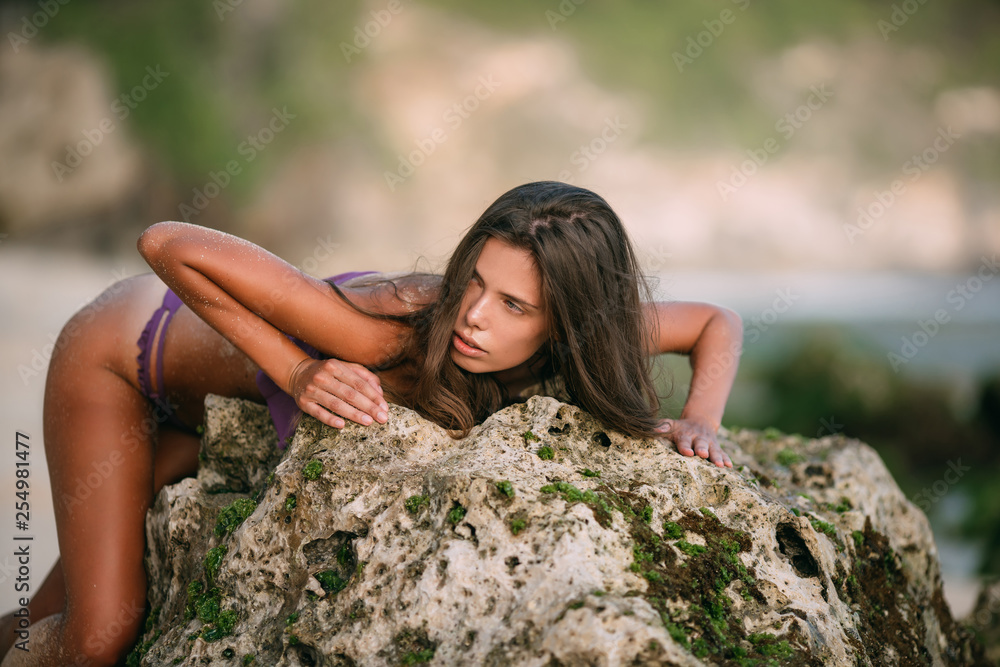 Sexy woman in purple colour swimsuit posing on sandy beach with big stones on background Sexy woman in purple colour swimsuit posing on sandy beach with big stones on background