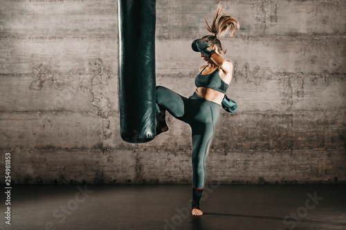 Dedykowana silna brunetka z kucykiem, w odzieży sportowej, boso i w rękawicach bokserskich kopiąc worek na siłowni.