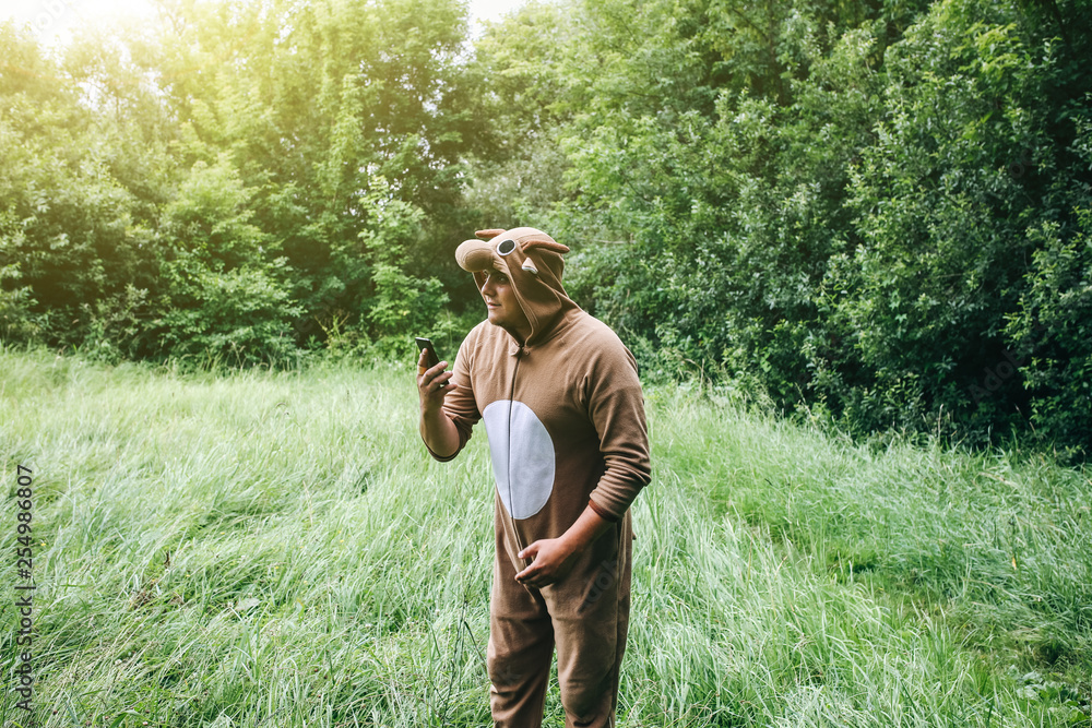 Young man is standing in the forest in cosplay costume of a cow. Guy in the  funny animal pyjamas sleepwear is holding smartphone in the nature.  Halloween ideas for party. Stock Photo |