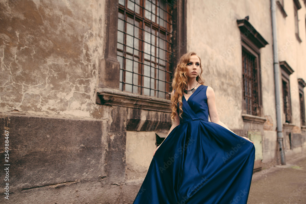 Young beautiful blonde woman in gorgeous blue long dress walking the street near the old building in the old city.