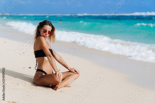 Beautiful girl in black swimsuit and sunglasses resting near ocean on beach