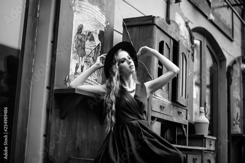 Gorgeous young woman in long blue dress posing in the outdoor cafe with the vintage exterior. Wooden furniture. Black and white shot