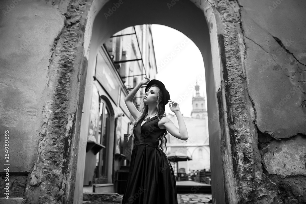 Black and white shot of beautiful blonde woman in gorgeous blue dress and hat walking under arch of the old building in the old city. Vintage shot concept.