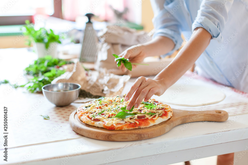 Woman is cooking pizza in cozy home kitchen. Female hands are decorating italian dinner with greens, fresh basil, arugula. Homemade pizza is served on wooden board on table. Lifestyle moment. Close up