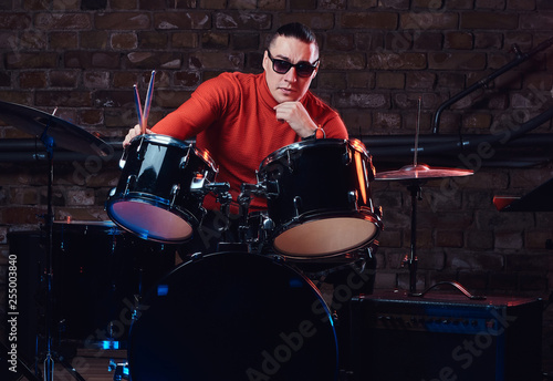 Young stylish musician in sunglasses sits behind the drum set against a brick wall, thoughtfully looking at the camera. Perform in a night club