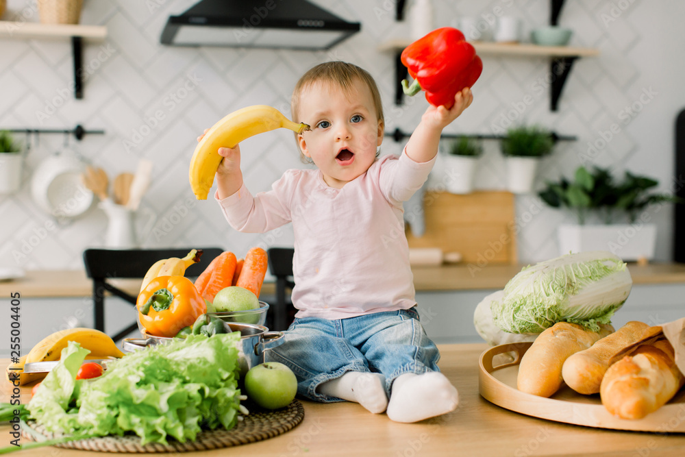 Cheerful baby child holds banana and red pepper while sitting on a table on the background of a modern light kitchen Cheerful baby child holds banana and red pepper while sitting on a table on the background of a modern light kitchen