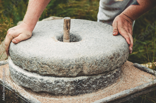 The ancient quern stone hand mill with grain. The man grinds the grain into flour with the help of a millstone. Men's hands on a millstone. Old grinding stones turned by hands photo