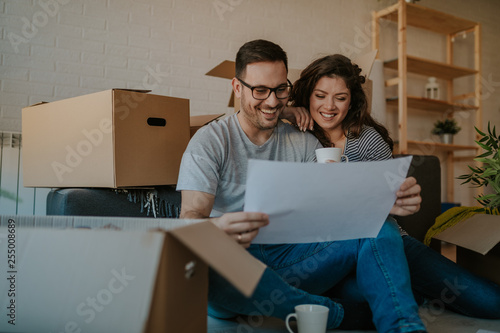 Beautiful young couple in casual clothes is discussing plan of their new house and smiling while sitting on the floor near boxes for move. Woman is drinking a coffee.