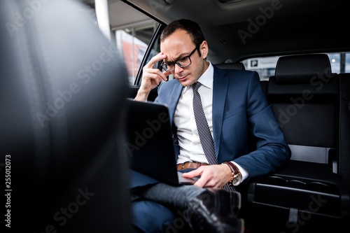 Brooding businessman with glasses sitting in the back seat of a car working on a laptop solving problems © pantovich