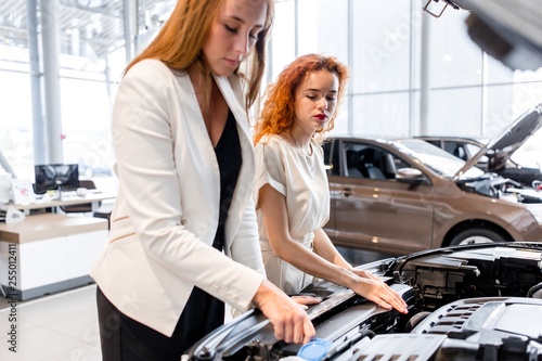 Two girls standing in front of an open car hood. Choosing a new car in the showroom © pantovich