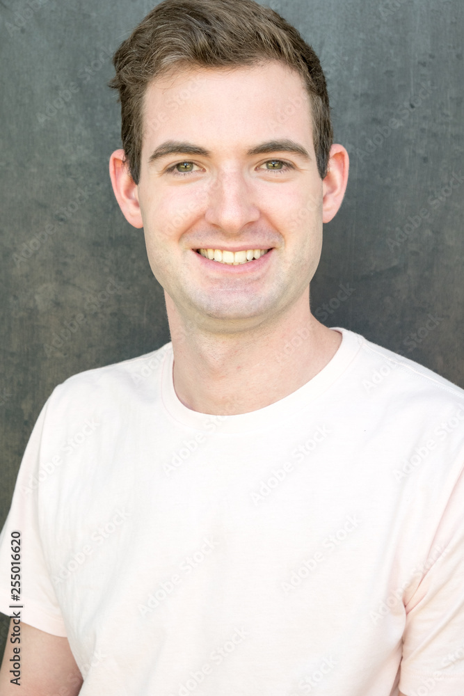 Portrait of a handsome man, Caucasian, in his 20s wearing a pale pink t-shirt against a textures grey background, looking at camera, smiling. Portrait of a handsome man, Caucasian, in his 20s wearing a pale pink t-shirt against a textures grey background, looking at camera, smiling.