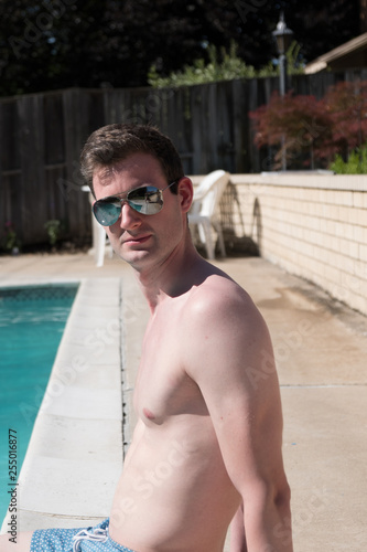 Portrait shot of healthy fit man sitting by the pool with feet in the water with no shirt and blue swim trunks in aviator sunglasses. White shirtless athletic model by outside backyard pool.