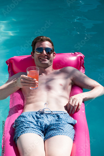 Trim young white man holding a drink and smiling on a bright pink raft in a swimming pool. Man smiling in swimming pool wearing aviator sunglasses. Relaxing in the swimming pool looking at camera.