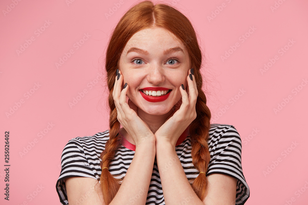 Portrait of amazed pretty red-haired girl with two braids keeps hands near her face and smiling lively with red lips, looking camera with widely opened happy blue eyes, isolated on pink background Portrait of amazed pretty red-haired girl with two braids keeps hands near her face and smiling lively with red lips, looking camera with widely opened happy blue eyes, isolated on pink background