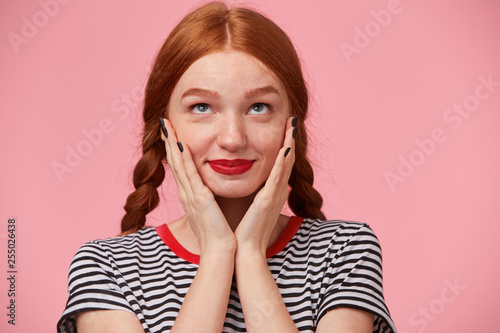 Close up of a pretty nice heartwarming red-haired girl with two braids keeps hands near her face and looking dreamingly with inspiration upwards, thinking about love, isolated on pink background