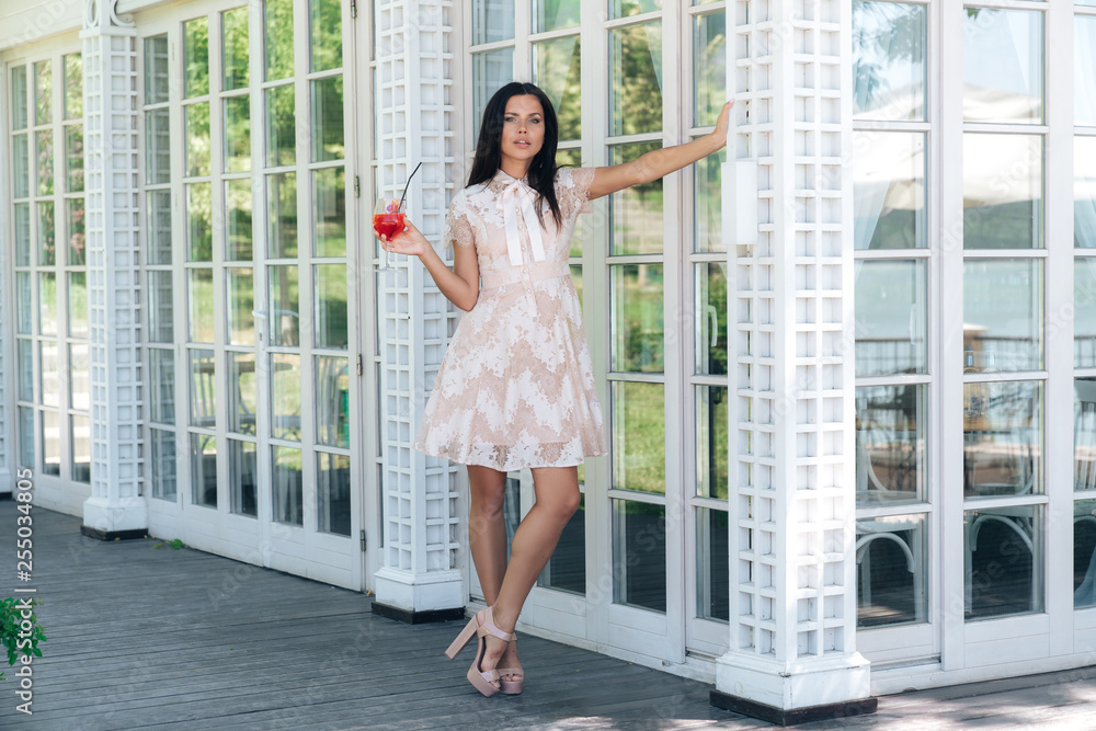 Beautiful brunette girl with glass of lemonade posing in beige colour dress outside cafe near a wooden and glass wall Beautiful brunette girl with glass of lemonade posing in beige colour dress outside cafe near a wooden and glass wall