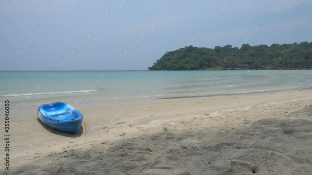 locked shot from clean white yellow sand beach to turquoise calm ocean on a sunny blue sky day, blue small boat ashore, no people, jungle shoreline on the far horizon. locked shot from clean white yellow sand beach to turquoise calm ocean on a sunny blue sky day, blue small boat ashore, no people, jungle shoreline on the far horizon.