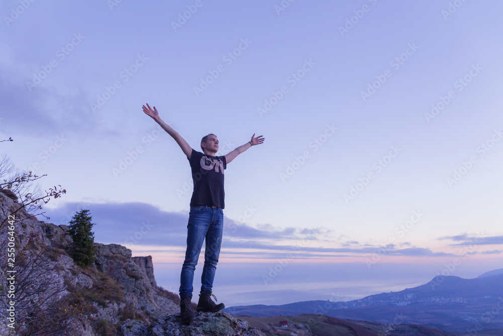 A young guy in a black shirt is standing on top of a mountain, spreading his arms toward the sky against the backdrop of a purple sunset.