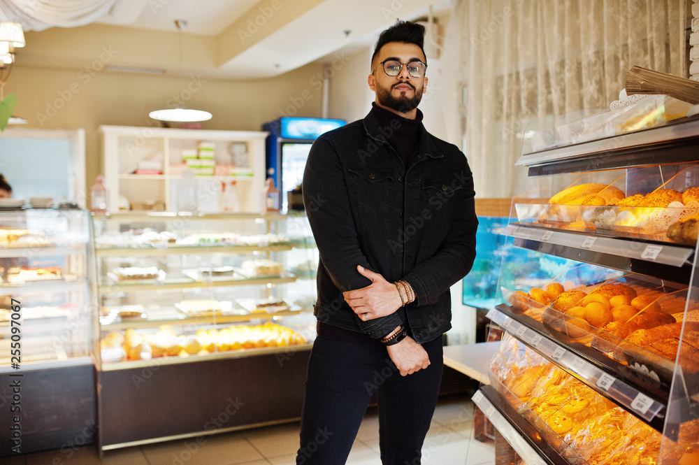 Arab man wear on black jeans jacket and eyeglasses in cafe choose bakery. Stylish and fashionable arabian model guy.