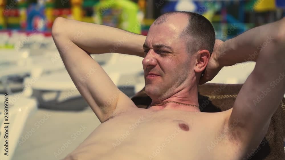 Close-up, Portrait: Bald Italian man is lying on the beach, on a sun lounger, sunbathing. Frowning from the sun, looking away. Close-up, Portrait: Bald Italian man is lying on the beach, on a sun lounger, sunbathing. Frowning from the sun, looking away.