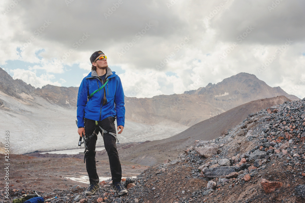 Obraz premium Portrait of a professional guide of a mountaineer in a cap and sunglasses with an ice ax in his hand against the background of the sky and the snow-capped mountains of the Caucasus
