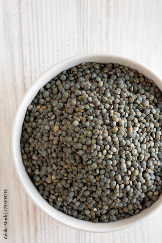 Dry green french lentils in gray bowl over white wooden surface, top view. Flat lay, overhead, from above. Close-up.