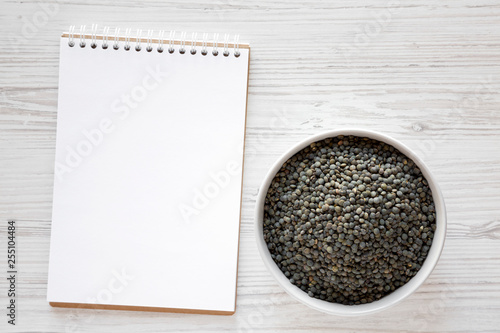 Green french lentils in gray bowl, blank notepad over white wooden background, top view. Flat lay, overhead, from above.