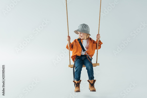 cute kid in jeans and orange shirt sitting on swing and looking away on grey background