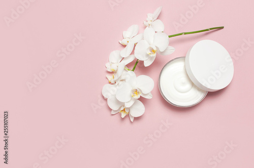 Beauty Spa concept. Opened plastic container with cream and White Phalaenopsis orchid flowers on pink background Flat lay top view. Herbal dermatology cosmetic hygienic cream, organic cosmetic Natural