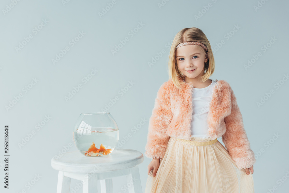 adorable kid in faux fur coat and skirt looking at camera and standing near to stool with fishbowl isolated on grey adorable kid in faux fur coat and skirt looking at camera and standing near to stool with fishbowl isolated on grey