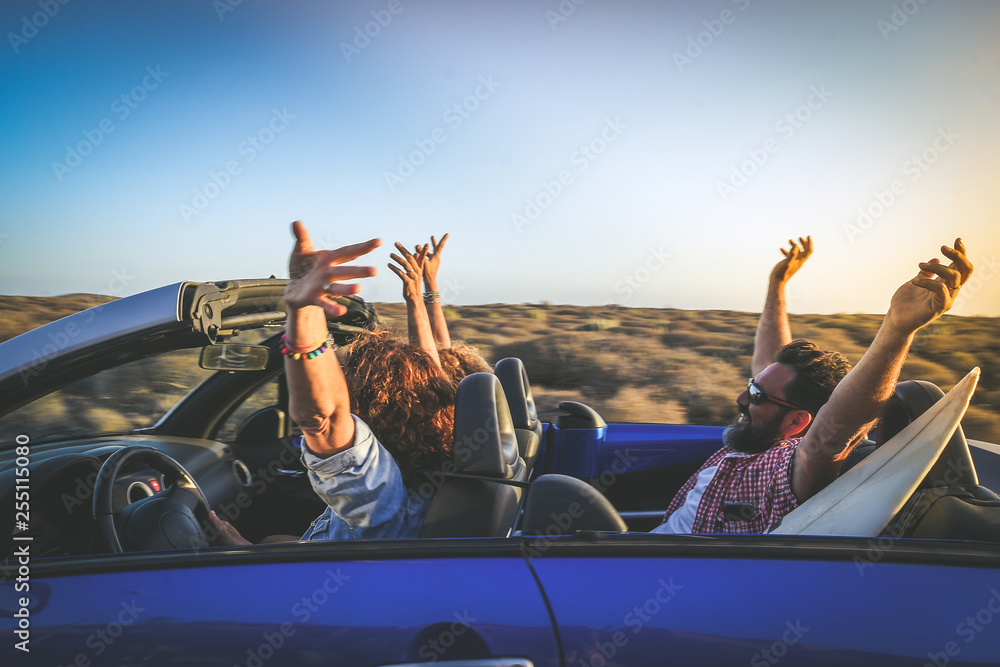 Side view group three euphoric friends in convertible car twisting and waving Two curly girls and charming bearded man on vacation having fun in topless auto Arms up palms open driving in the sunlight - obrazy, fototapety, plakaty 