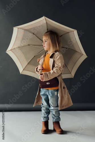 serious child in trench coat and jeans holding umbrella and looking away