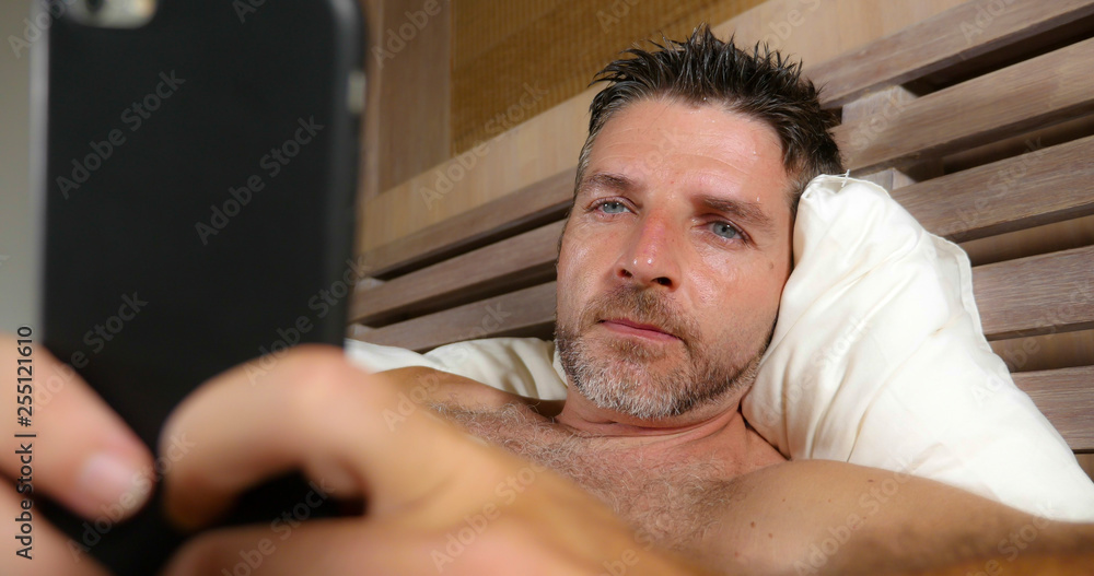 Obraz premium lifestyle portrait of young attractive and happy man lying on bed at home bedroom using mobile phone enjoying internet social media app or online gambling and dating