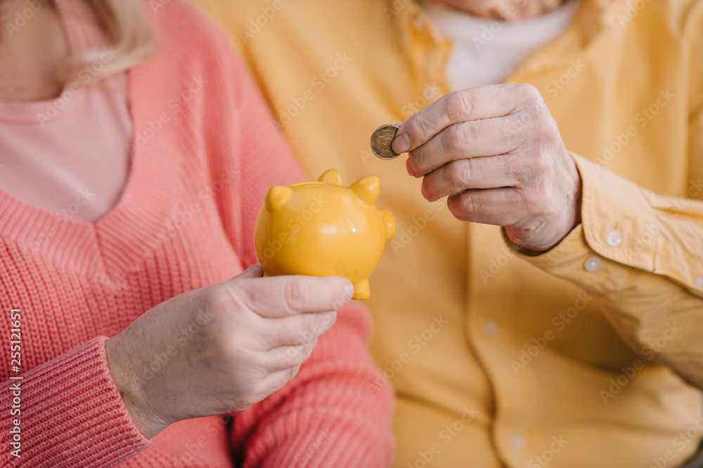 cropped view of senior couple putting coin in yellow piggy bank