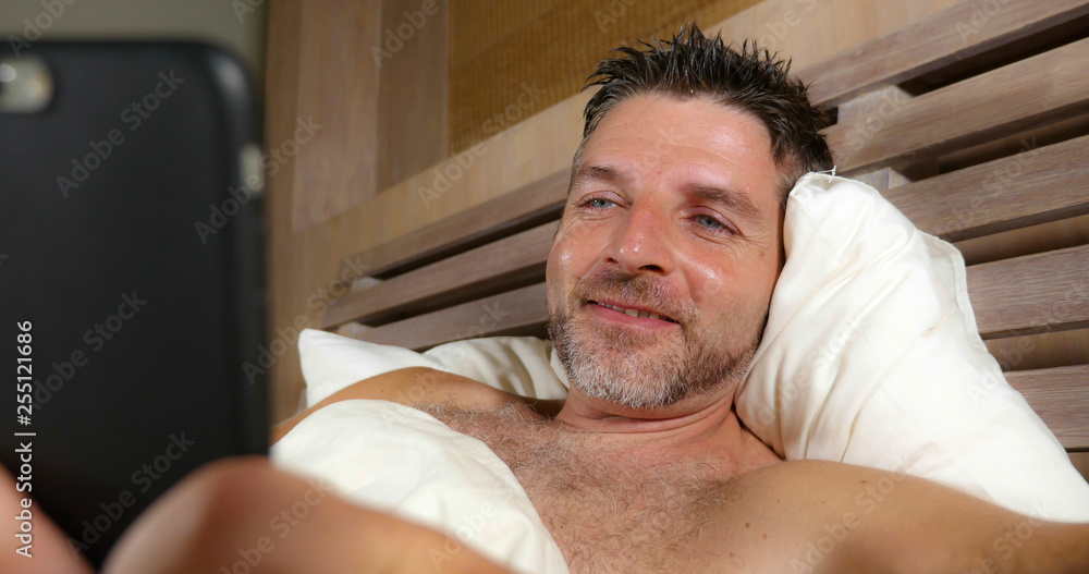 Fototapeta premium lifestyle portrait of young attractive and happy man lying on bed at home bedroom using mobile phone enjoying internet social media app or online gambling and dating