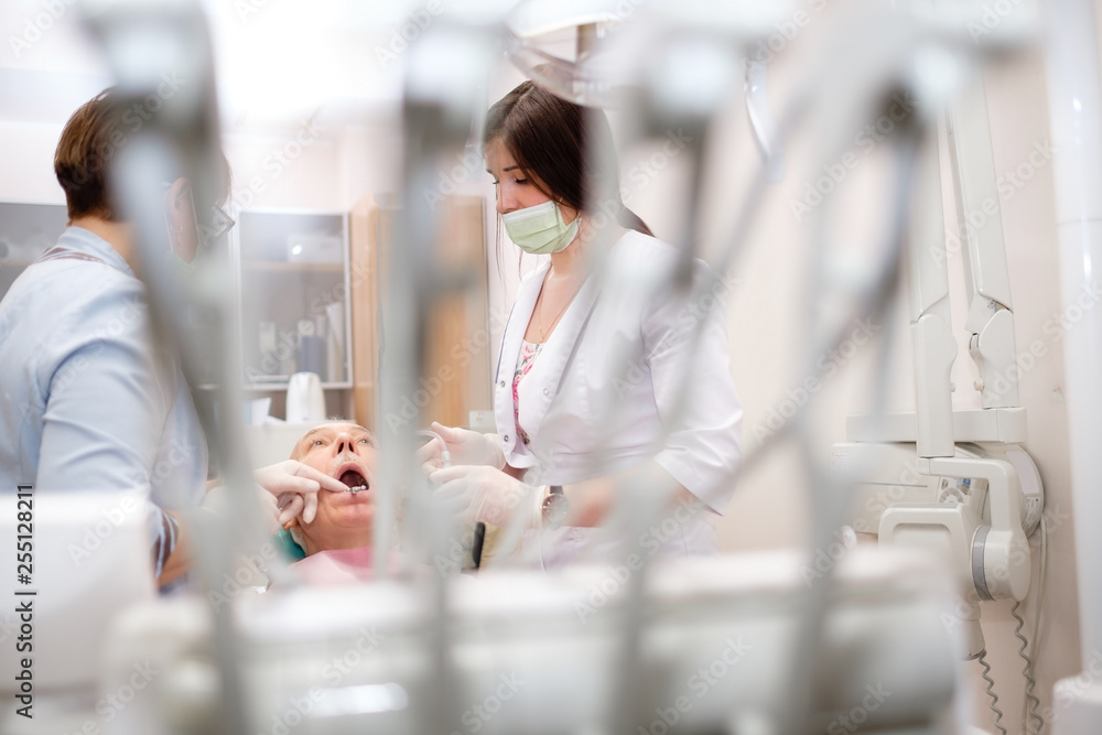Modern stomatology private clinic with professional equipment. Two women dentists working with senior man patient. Dental care for elder people. Dentist holding dental device. Modern stomatology private clinic with professional equipment. Two women dentists working with senior man patient. Dental care for elder people. Dentist holding dental device.