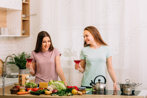 Healthy eating. Dieting together. Two young females in kitchen with fresh vegetables assortment. Red wine in glasses.