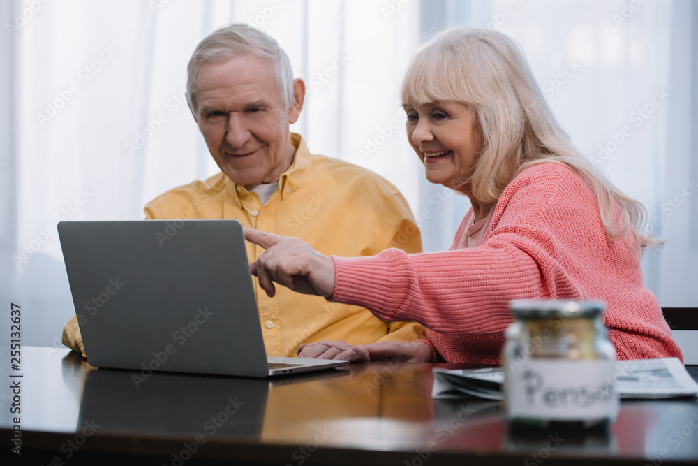 smiling senior couple sitting at table, pointing with finger and using laptop smiling senior couple sitting at table, pointing with finger and using laptop