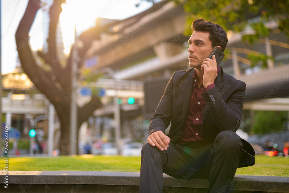 Hispanic businessman thinking while talking on phone with nature in city Hispanic businessman thinking while talking on phone with nature in city