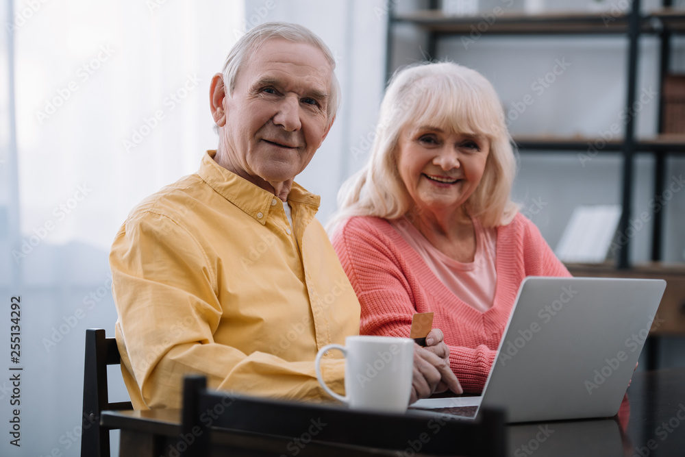 senior couple looking at camera and using laptop while sitting at table with coffee cup at home senior couple looking at camera and using laptop while sitting at table with coffee cup at home