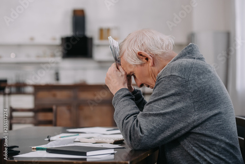 stressed senior man in casual clothes sitting at table with paperwork and holding money at home