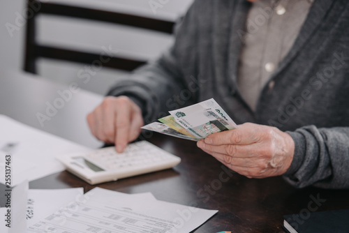 cropped view of senior man sitting at table with paperwork and using calculator while counting money