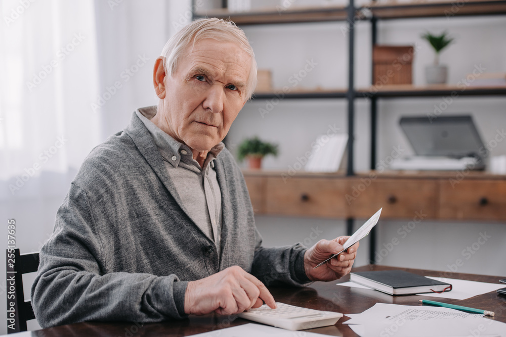 senior man sitting at table with paperwork, looking at camera and using calculator while counting money senior man sitting at table with paperwork, looking at camera and using calculator while counting money