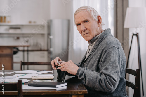 male pensioner sitting at table with paperwork, looking at camera and putting money in wallet
