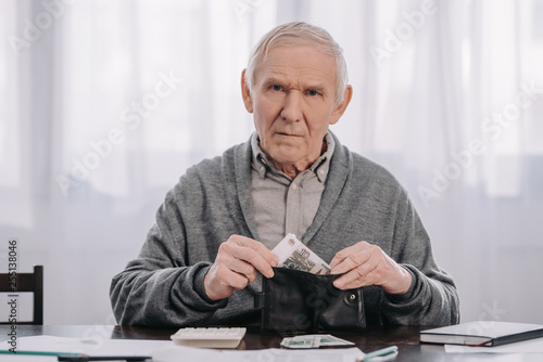 male pensioner sitting at table and putting money in wallet while looking at camera