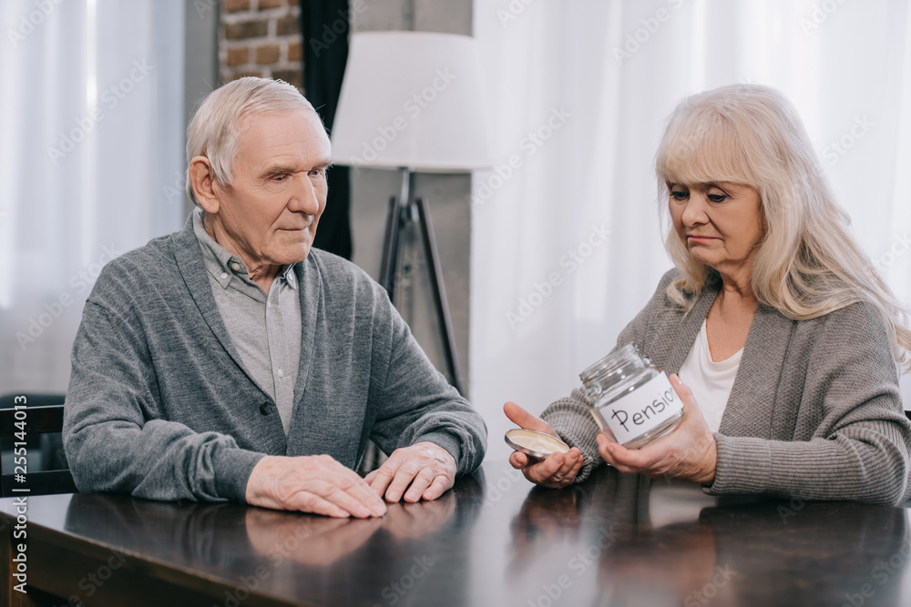 upset senior couple sitting at table and holding empty glass jar with 'pension' lettering upset senior couple sitting at table and holding empty glass jar with 'pension' lettering